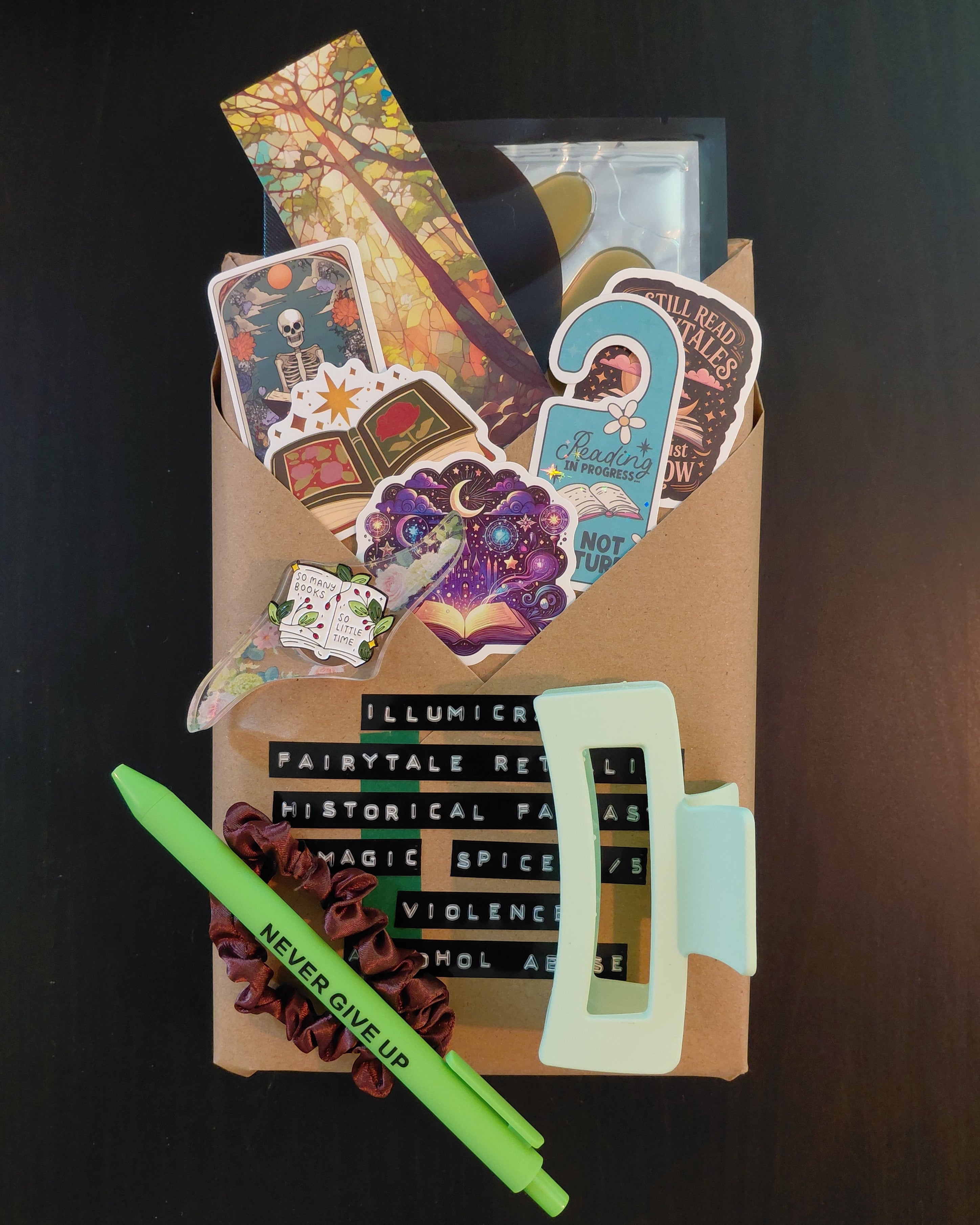 A curated flat-lay of a themed gift box or subscription package on a dark wood background. The package contains a variety of colorful stickers, a light green hair clip, a green pen with 'NEVER GIVE UP' printed on it, a floral printed thumb page holder, a bookmark, small brown scrunchie, eye mask, enamel pin and an Illumicrate Book wrapped in Brown Paper.